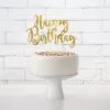 Златен сладък топер с надпис "Happy Birthday" върху бял торта със цветни сладки върху фон на бял тухлен стена, за рожден ден, парти декорация, празненство, торта за рождения ден, Party Deluxe.