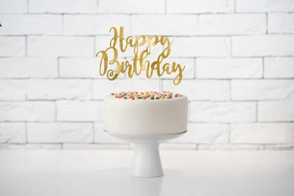 Златен сладък топер с надпис "Happy Birthday" върху бял торта със цветни сладки върху фон на бял тухлен стена, за рожден ден, парти декорация, празненство, торта за рождения ден, Party Deluxe.