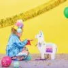 Beautiful girl in colorful costume playing with paper unicorn piñata at vibrant birthday party, decorated with balloons and golden streamers, cheerful celebration atmosphere.