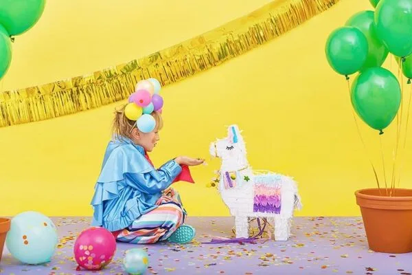 Beautiful girl in colorful costume playing with paper unicorn piñata at vibrant birthday party, decorated with balloons and golden streamers, cheerful celebration atmosphere.