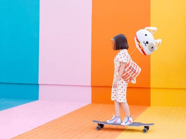 Радостна млада момиче със зайче балон, носеща раница, караща скейтборд пред цветна фонова стена за парти и забавление с събития на Party Deluxe.