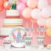 Pink birthday cake with unicorn and rainbow decorations, party cups with unicorn toppers, balloons in the background, perfect for kids' celebrations and themed parties.