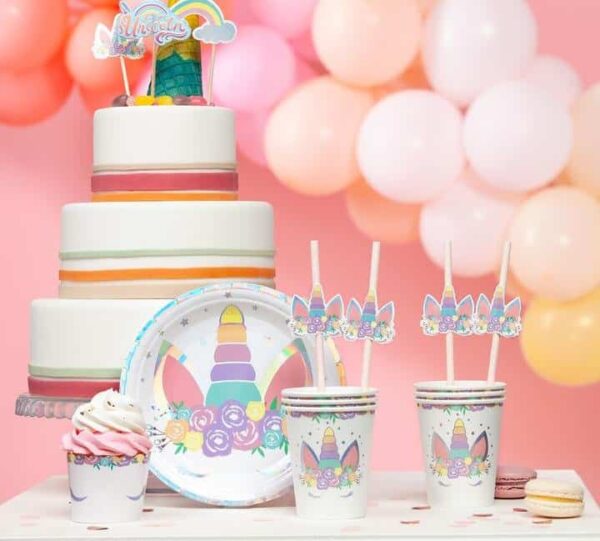 Pink birthday cake with unicorn and rainbow decorations, party cups with unicorn toppers, balloons in the background, perfect for kids' celebrations and themed parties.