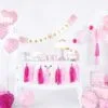 Pink baby girl celebration decorations with balloons, banner, cake, and gifts for a baby shower or first birthday party.