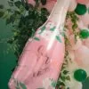 Бутилка балон с надпис „Cheers to you“ за празненства, украсена с цветя и зелени листа, на фона на балони в розово, зелено и бяло, перфектна за парти и събития.