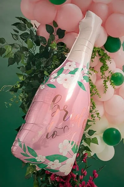 Бутилка балон с надпис „Cheers to you“ за празненства, украсена с цветя и зелени листа, на фона на балони в розово, зелено и бяло, перфектна за парти и събития.