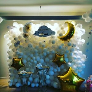 Разделена декорация за детско парти с балони и фигури, подходящи за празнуване на бебе или новородено, с бял фон и златисти ивици и звездички за стилна конфигурация.