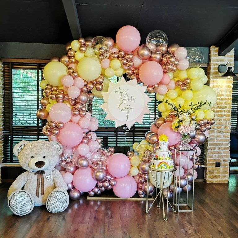 Розова и златист балонна декорация за рожден ден с голямо поздравително табло, плюшен мечок и цветна торта, идеална за празненство, организирана от Party Deluxe.